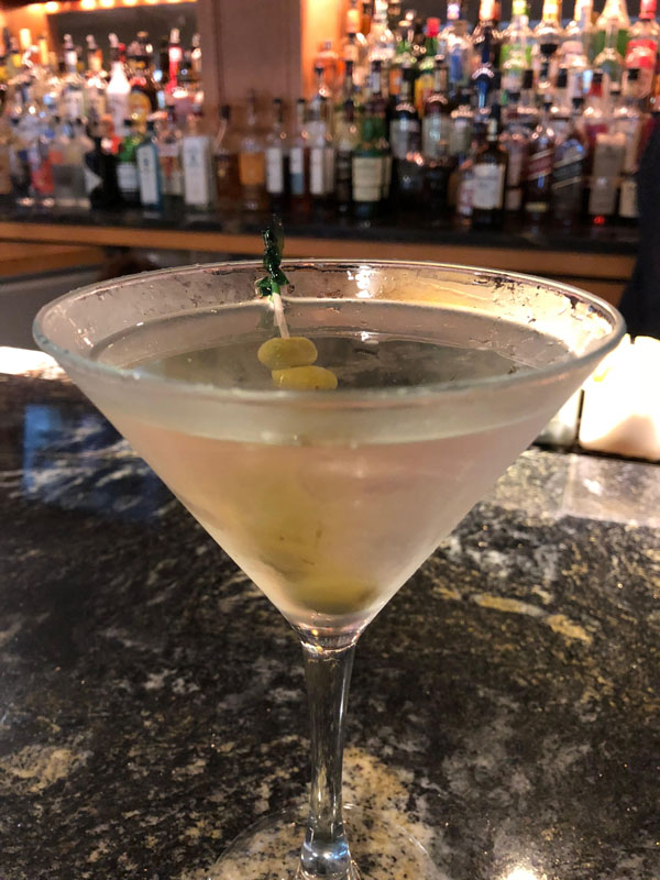Martini, on a dark bar, with liquor bottles in the background
